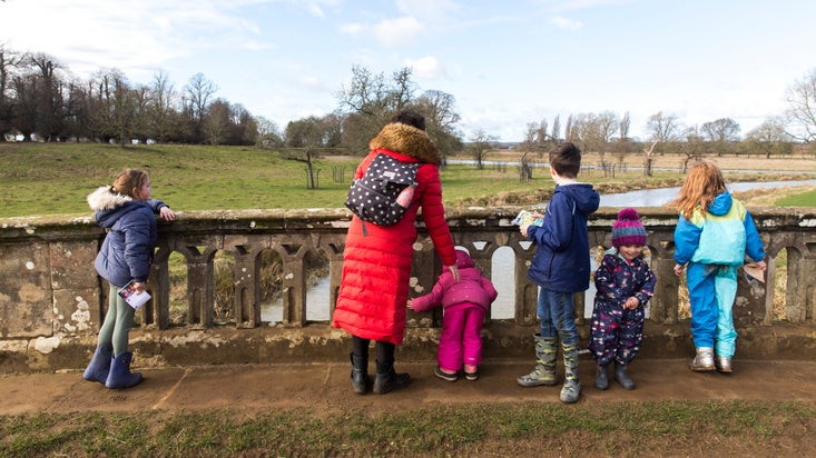 Family visitors in colourful coats, looking out over the old stone bridge at the river Dene, with a wintry view of the parkland in the distance.